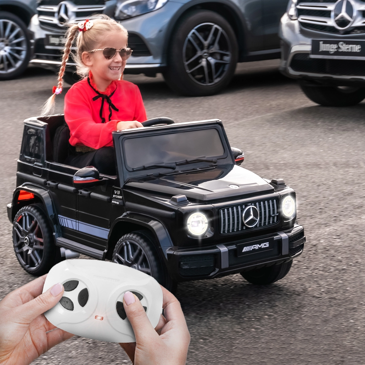 2,4 GHz-Fernbedienung mit First-Funktion Mädchen steuert das Kinderauto über einen Parkplatz, Fernbedienung in Hand eines Erwachsenen